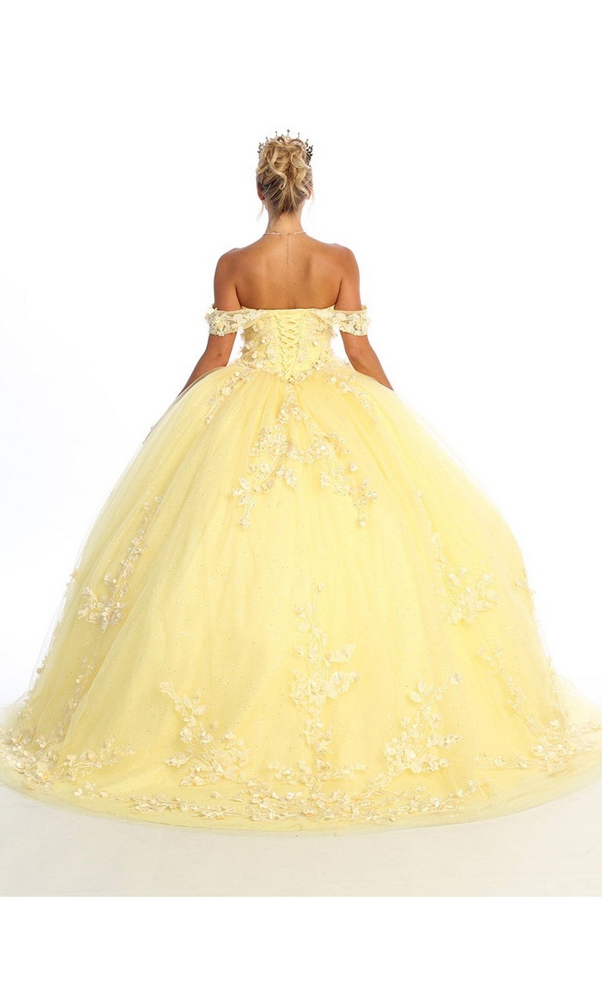 A person stands facing away, wearing the Quinceanera Dress CHF3074 By Chicas—a yellow off-shoulder ball gown featuring floral appliques and delicate lace embroidery—paired with a tiara, set against a plain white background.